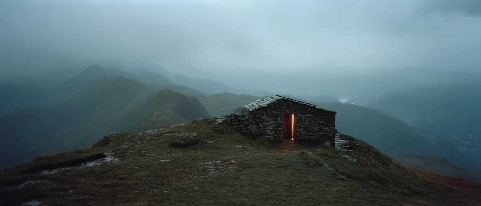 A small stone shelter on an exposed ridge in storm cloud, light showing in its doorway