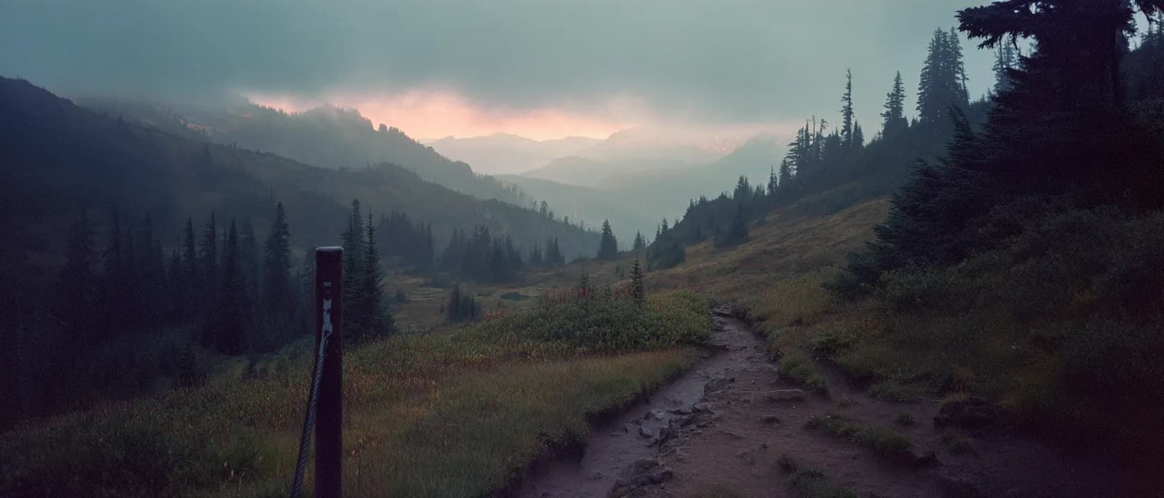 A dirt trail leading from a marker post into a misty mountain valley at first light